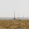 Campo árido con vegetación baja y varias torres de perforación de petróleo no convencional alineadas en el horizonte, algunas con llamas visibles, bajo un cielo despejado, en Añelo (Argentina) en la zona del yacimiento de Vaca Muerta.