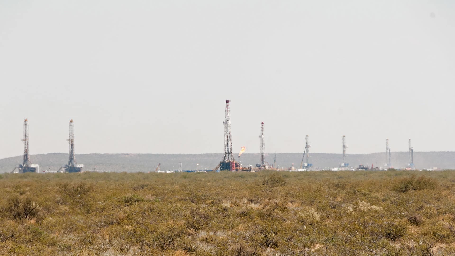 Campo árido con vegetación baja y varias torres de perforación de petróleo no convencional alineadas en el horizonte, algunas con llamas visibles, bajo un cielo despejado, en Añelo (Argentina) en la zona del yacimiento de Vaca Muerta.