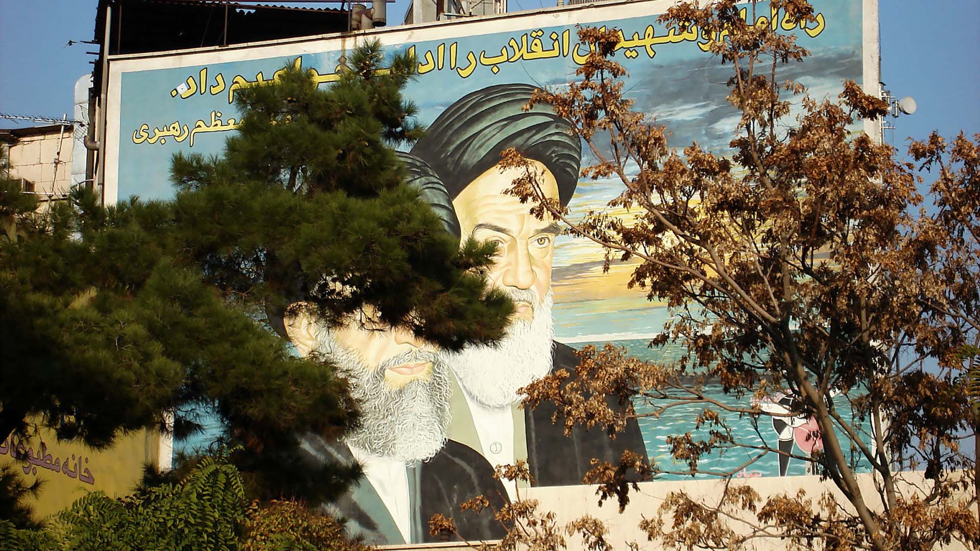 Valla pintada con los rostros de los ayatolás Jomeini y Jamenei en un edificio en Teherán (Irán). La valla está cubierta parcialmente a la izquierda y a la derecha con árboles. La foto data de 2006.