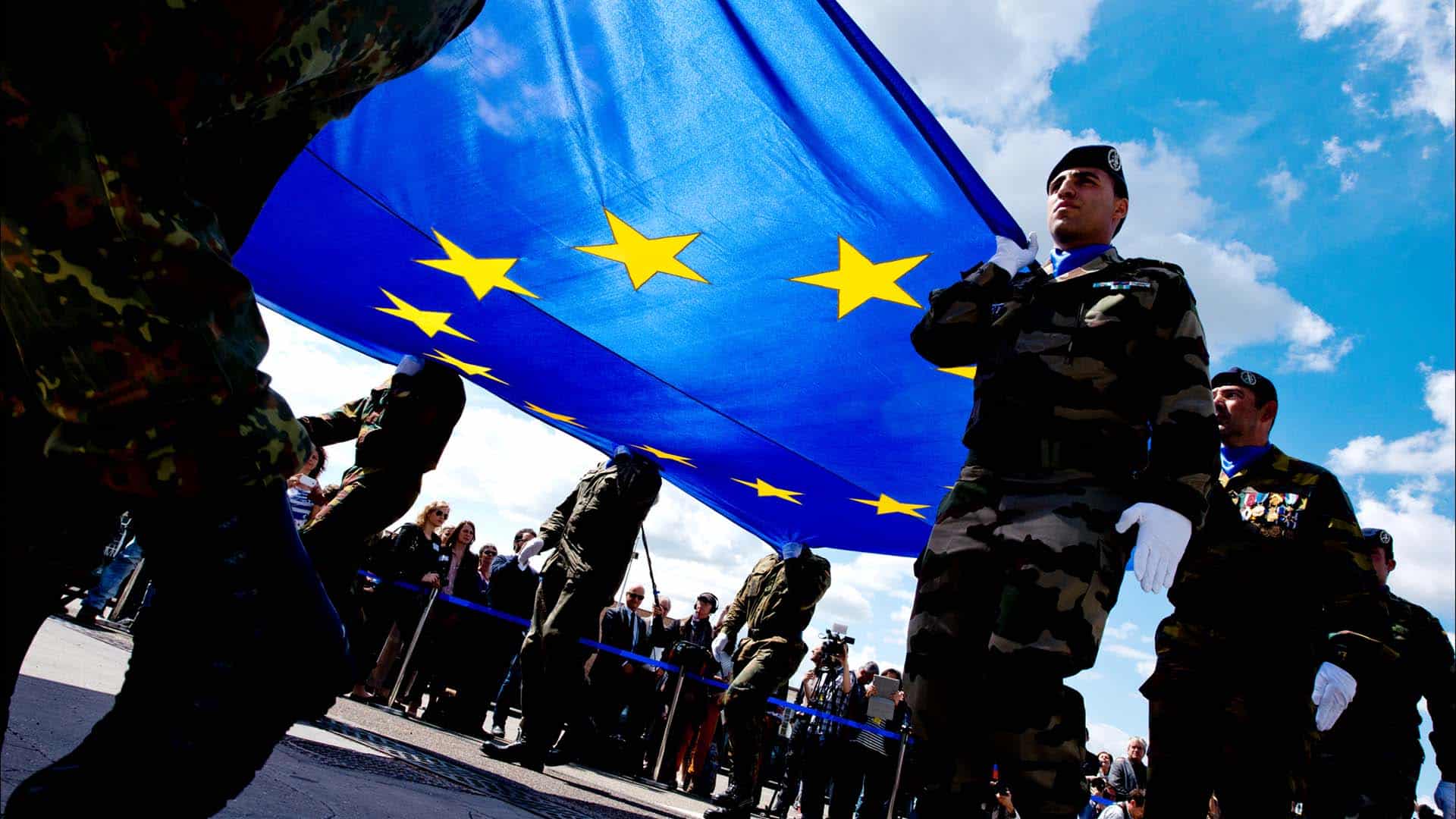 Grupo de militares sosteniendo una gran bandera azul con estrellas amarillas, símbolo de la Unión Europea, durante una ceremonia al aire libre frente al Parlamento Europeo, en la primera sesión plenaria del Parlamento Europeo que marca el inicio para el mandato 2014-2019. Al fondo el cielo parcialmente nublado y una multitud tras una valla observando.