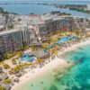 Vista aérea de una playa turística de arena blanca y aguas turquesas en Cancún con grandes hoteles, junto a una laguna y áreas urbanizadas.