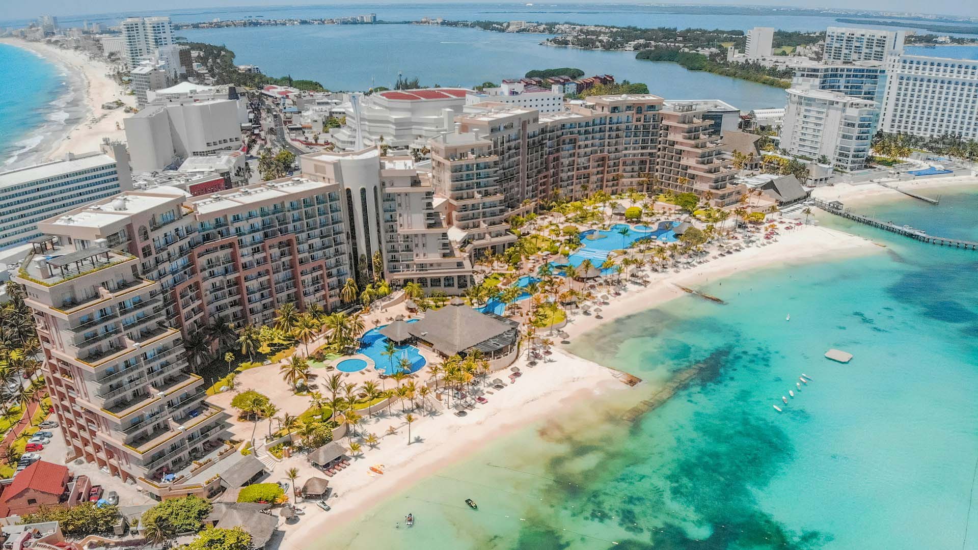 Vista aérea de una playa turística de arena blanca y aguas turquesas en Cancún con grandes hoteles, junto a una laguna y áreas urbanizadas.
