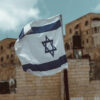 Bandera de Israel ondeando en primer plano. Al fondo (desenfocado), paredes y muros de edificaciones de la ciudad de Jerusalén, tanques de agua, antenas parabólicas y palmeras.