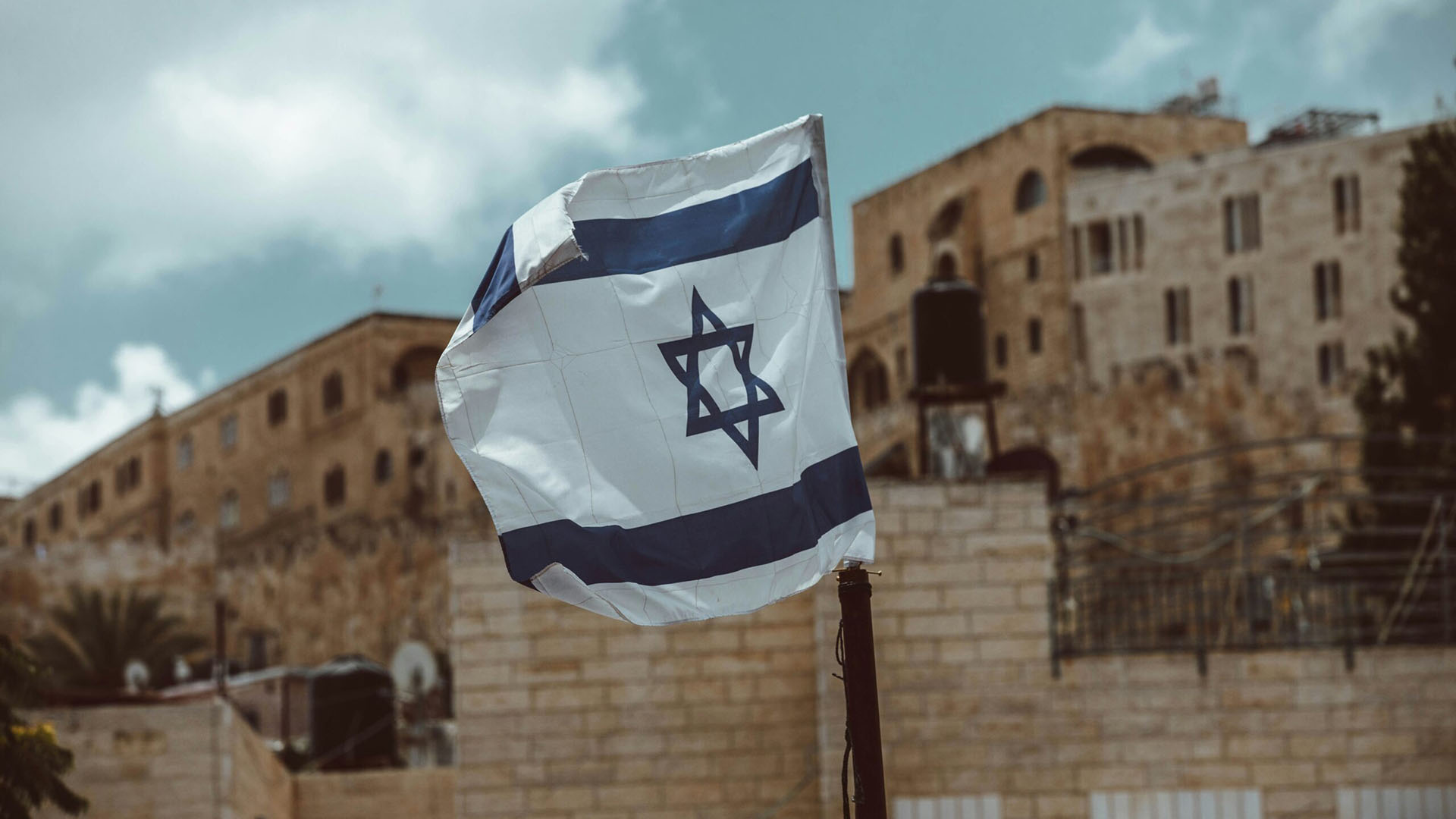 Bandera de Israel ondeando en primer plano. Al fondo (desenfocado), paredes y muros de edificaciones de la ciudad de Jerusalén, tanques de agua, antenas parabólicas y palmeras.