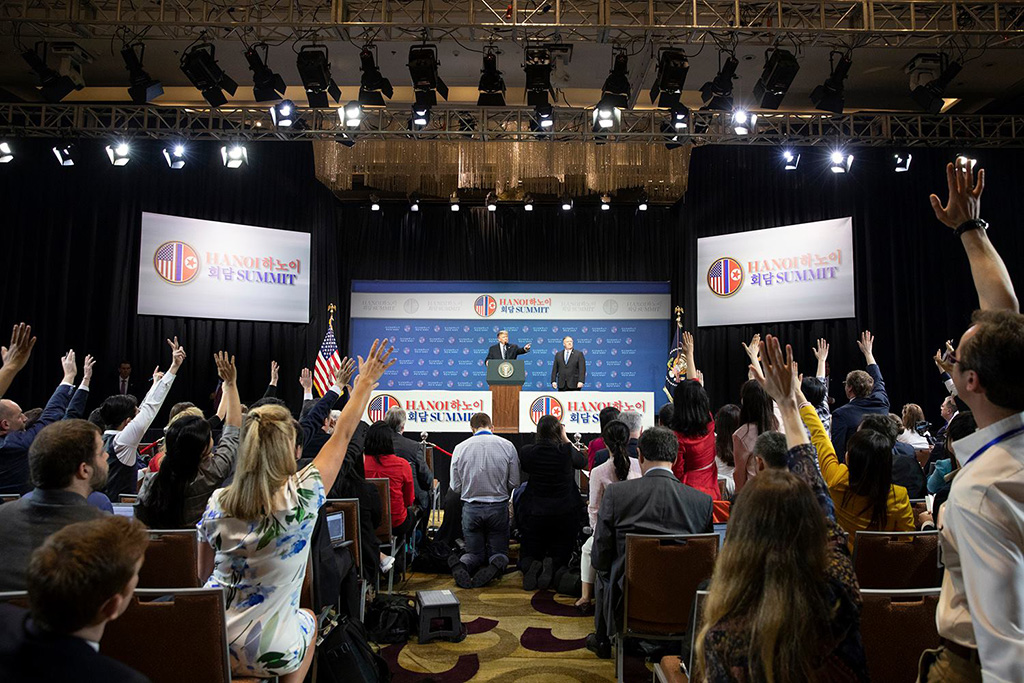 Periodistas levantan la mano en una sala de prensa en Hanói con Donald Trump en segundo plano.