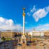 Vista de la Plaza de la Independencia y monumento a los fundadores en Kyiv, Ucrania, rodeada de edificios, torres, árboles y calles con coches y autobuses con gente al fondo