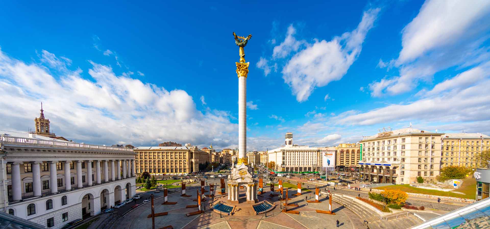 Vista de la Plaza de la Independencia y monumento a los fundadores en Kyiv, Ucrania, rodeada de edificios, torres, árboles y calles con coches y autobuses con gente al fondo