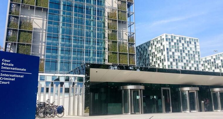 Headquarters of the International Criminal Court (ICC) in The Hague. In the left foreground, a blue sign with the ICC logo. In the background, a bicycle parking area and the visitors’ entrance to the building