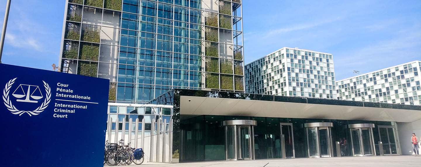 Headquarters of the International Criminal Court (ICC) in The Hague. In the left foreground, a blue sign with the ICC logo. In the background, a bicycle parking area and the visitors’ entrance to the building
