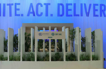 Detalle del pabellón de la Casa de Comercio de la COP28 en Dubái, Emiratos Árabes Unidos. Fondo: una suave cortina azul con las palabras “UNITE. ACT. DELIVER”. Primer plano: un escenario con plantas y un gran cartel blanco con los logotipos impresos de la CMNUCC y la COP28 EAU.