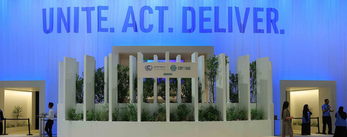 Detail of the COP28’s Trade House pavilion in Dubai, United Arab Emirates. Background: a soft blue curtain with the words “UNITE. ACT. DELIVER”. Foreground: a stage with plants and a big white poster with the printed logos of the UNFCC and the COP28 UAE