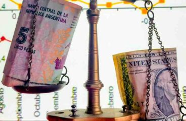 Detail of a balance scale holding Argentine pesos and dollars in their saucers. Background: Screen with timeline of the rise and fall of the stock market.
