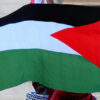 Persona sosteniendo la bandera palestina en la calle. Fondo: Facha de edificio a la luz del día.