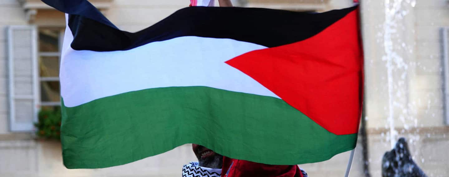 Persona sosteniendo la bandera palestina en la calle. Fondo: Facha de edificio a la luz del día.