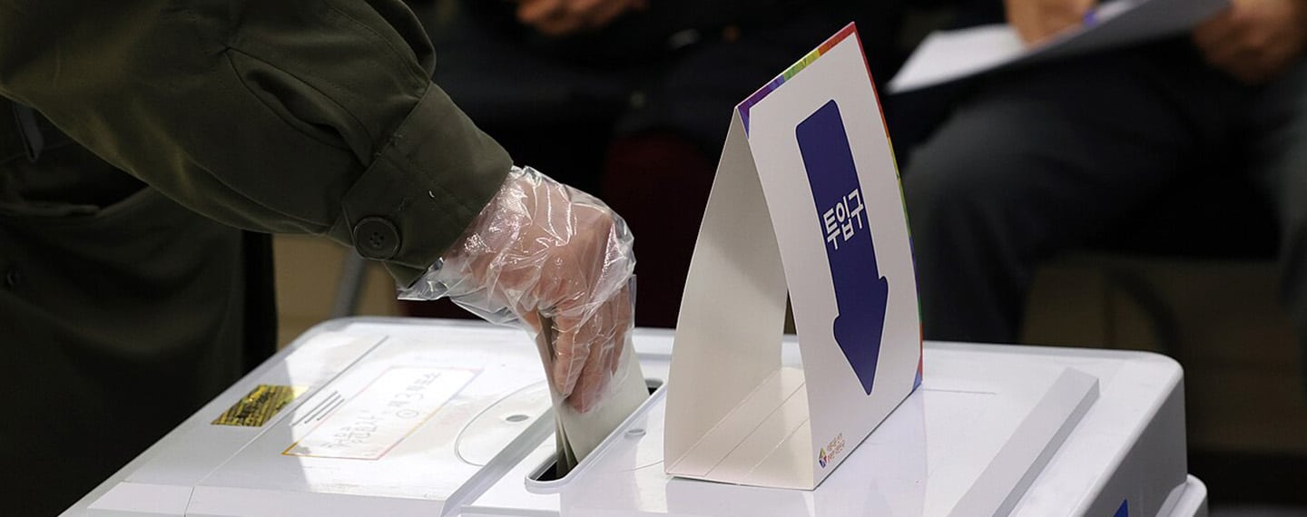 Persona ejerciendo su derecho al voto en un centro de votación. Primer plano: Mano introduciendo papeleta en una urna blanca.