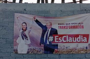Claudia Sheinbaum and Andrés Manuel López Obrador on a MORENA electoral propaganda poster in Dolores Hidalgo, Guanajuato (Mexico). The poster is hung on a cement block wall painted grey and yellow. At the top of the wall protrude pieces of wood with a barbed wire fence