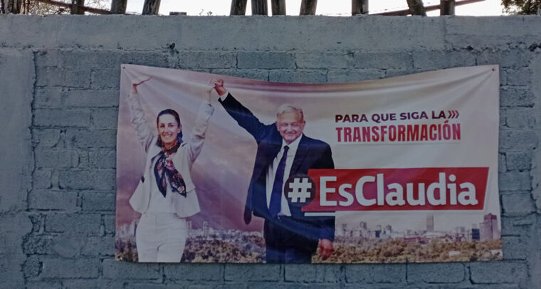 Claudia Sheinbaum y Andrés Manuel López Obrador en un cartel de propaganda electoral de MORENA en Dolores Hidalgo, Guanajuato (México). El cartel está colgado en una pared de bloques de cemento pintada de gris y amarillo. En la parte superior de la pared sobresalen trozos de madera con una alambrada