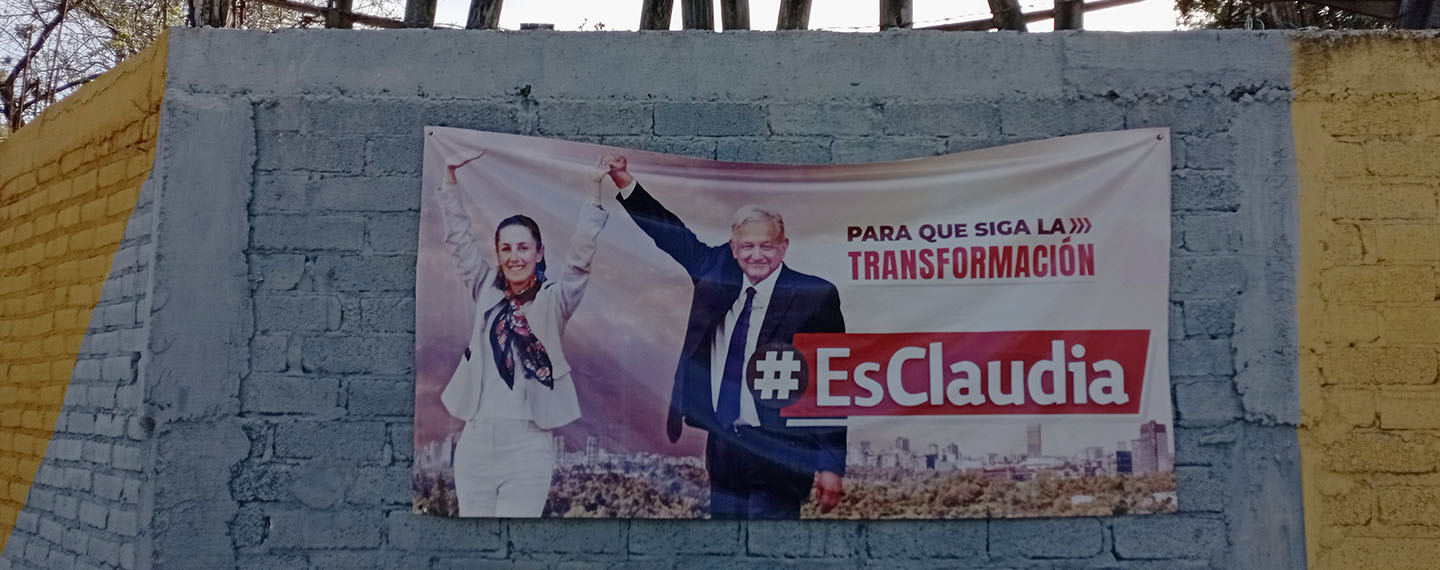 Claudia Sheinbaum and Andrés Manuel López Obrador on a MORENA electoral propaganda poster in Dolores Hidalgo, Guanajuato (Mexico). The poster is hung on a cement block wall painted grey and yellow. At the top of the wall protrude pieces of wood with a barbed wire fence