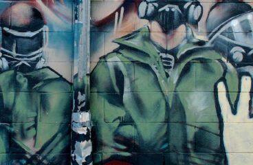 Graffiti detrás de Bloor West, cerca de la estación de metro Dundas West en Toronto (Canadá). El graffiti muestra en primer plan unas manos levantadas haciendo el símbolo de paz contra el fondo donde hay tres figuras enmascaradas que representan fuerzas militares y policiales y a la izquierda un rostro difuminado expresando un grito de ira