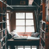 Interior de una librería con estructura de madera con niveles. Al fondo, un ventanal con unos cojines cómodos en el suelo y los lados estanterías llenas de libros