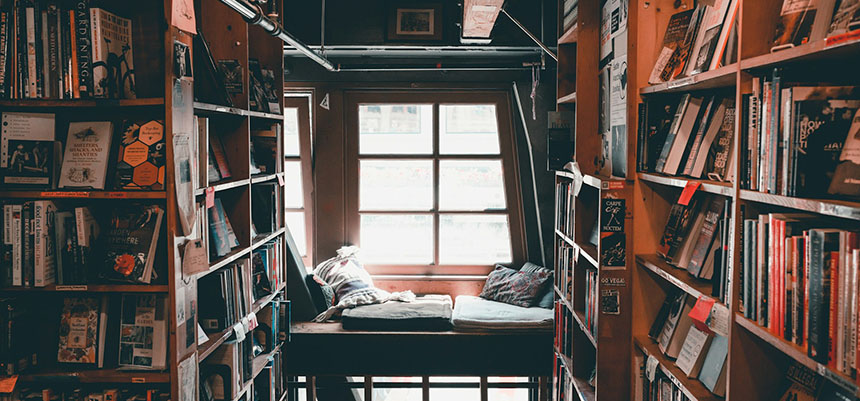 Interior de una librería con estructura de madera con niveles. Al fondo, un ventanal con unos cojines cómodos en el suelo y los lados estanterías llenas de libros