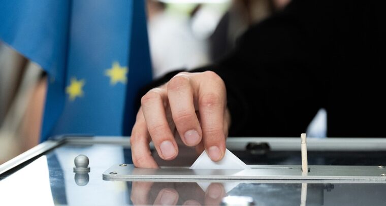 Persona depositando su voto en una urna. Fondo: Bandera de la Unión Europea.
