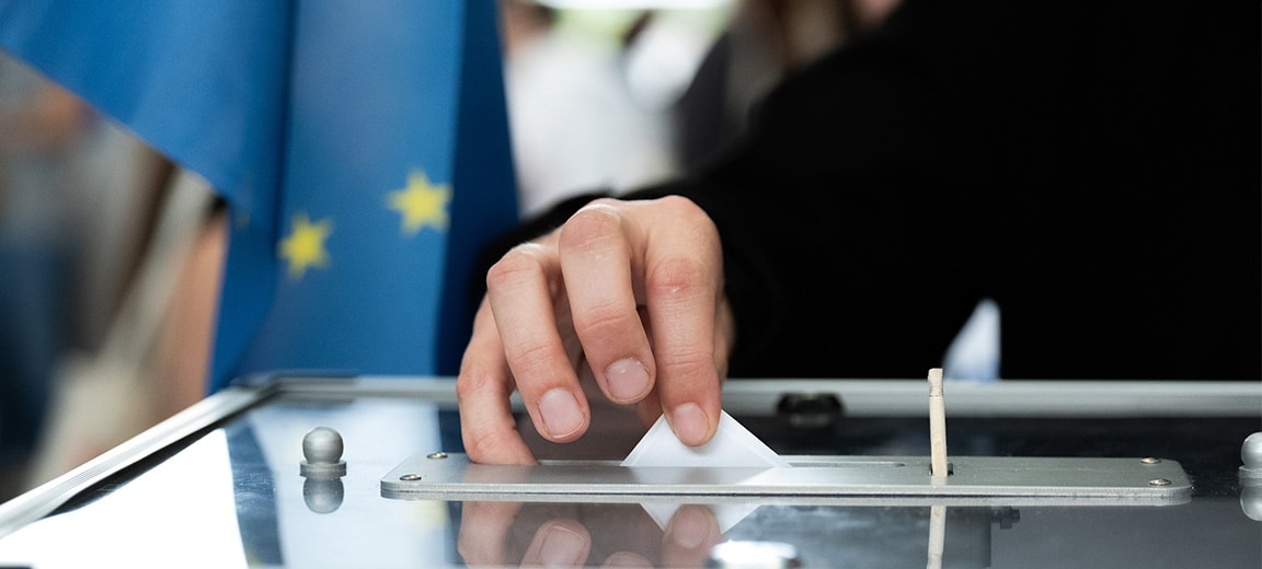 Persona depositando su voto en una urna. Fondo: Bandera de la Unión Europea.