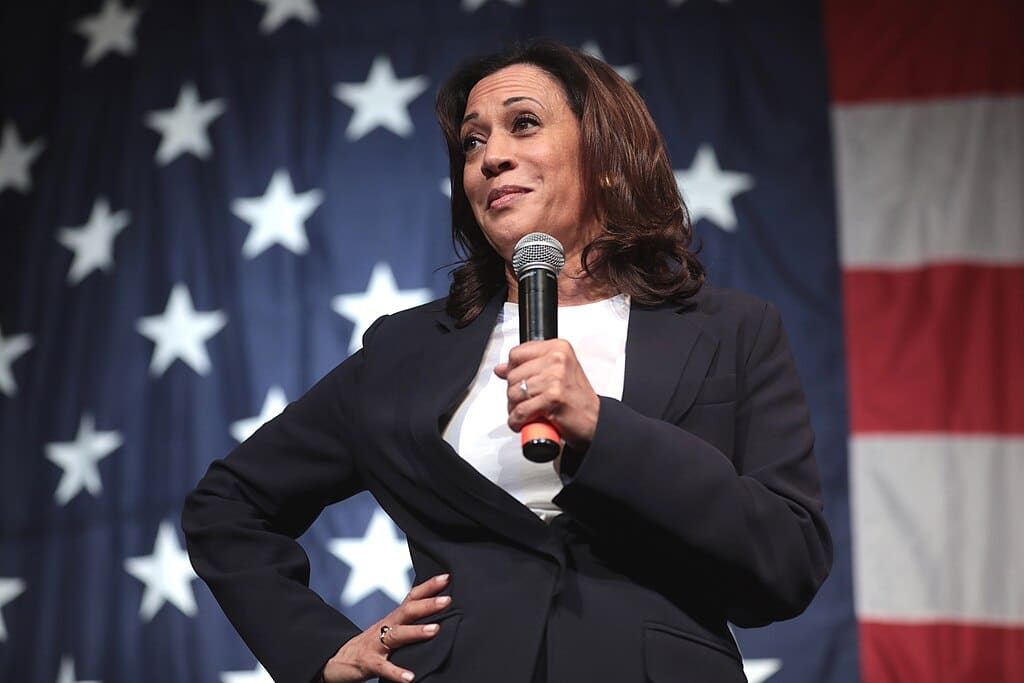 Kamala Harris, en aquel entonces Senadora, con traje azul marino y camisa blanca, de pie con la mano derecha en la cintura y con el micrófono en la derecha, y con una bandera de EEUU al fondo. Habla con los asistentes al Iowa Democratic Wing Ding 2019 en Surf Ballroom en Clear Lake (Iowa). Foto: Gage Skidmore (CC BY-SA 4.0)