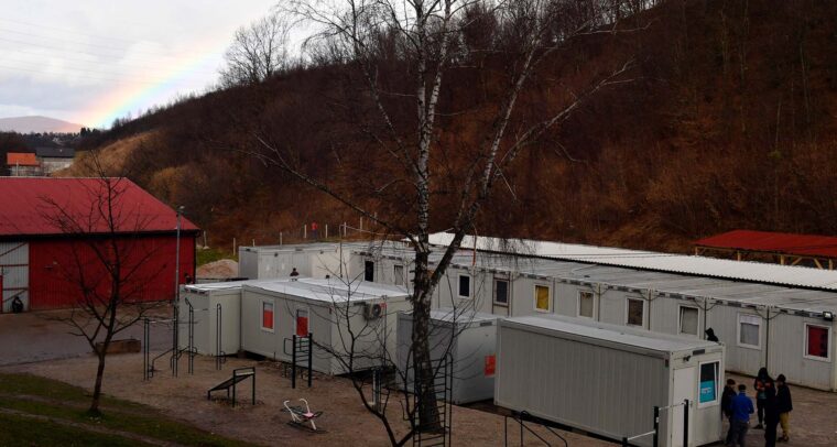 Centro de acogida de inmigrantes en Blazuj en Bosnia y Herzegovina, conformado por unas casetas prefabricadas en un terreno yermo en la ladera de una colina, con un pequeño parque infantil y un galpón de almacenamiento de color rojo a la izquierda. En el parque hay dos árboles sin hojas