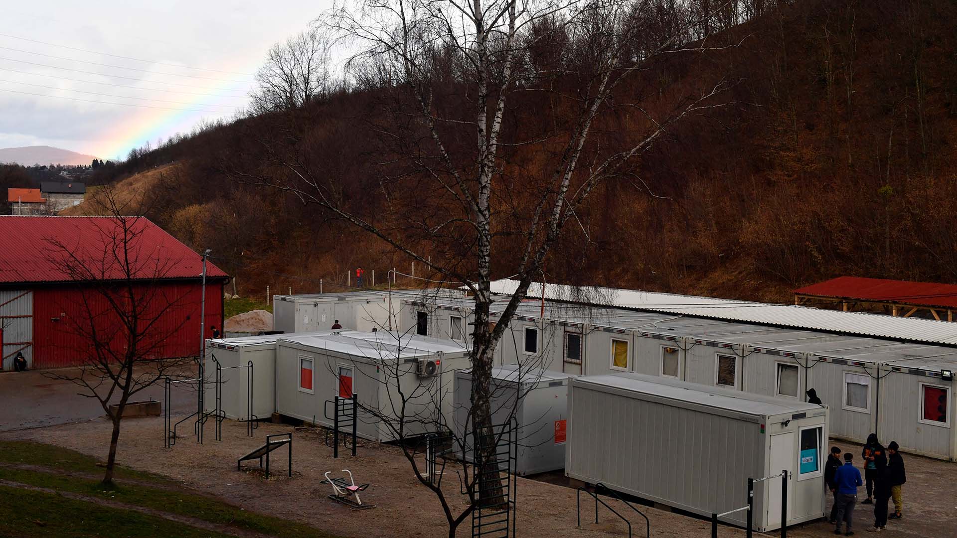 Centro de acogida de inmigrantes en Blazuj en Bosnia y Herzegovina, conformado por unas casetas prefabricadas en un terreno yermo en la ladera de una colina, con un pequeño parque infantil y un galpón de almacenamiento de color rojo a la izquierda. En el parque hay dos árboles sin hojas