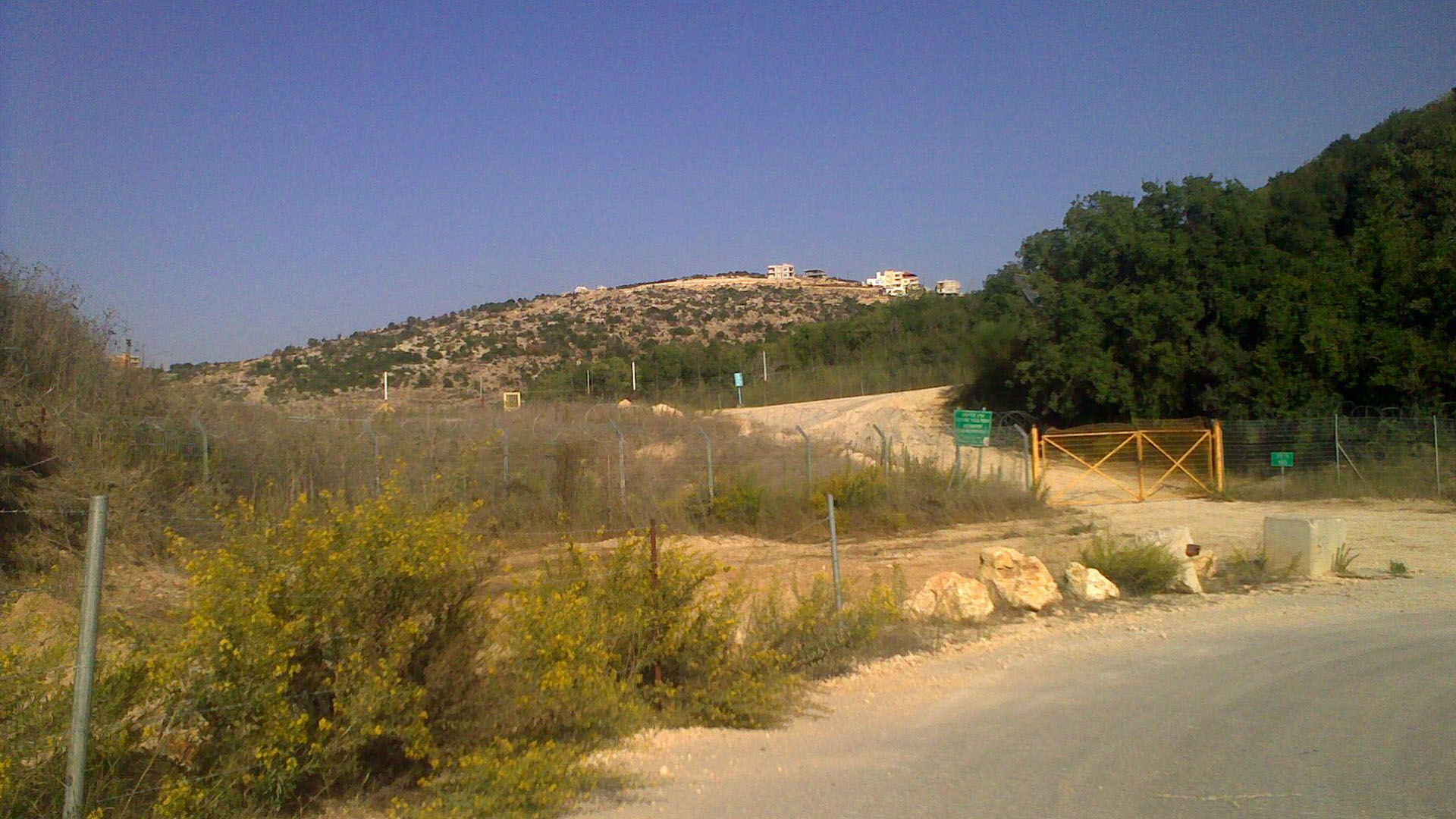 Punto 105 de la frontera entre Israel y Líbano. Al frente una verja de alambre electrificada carteles de señalización de color verde con letras blancas (ilegibles). Tras la verja una carretera de tierra conduce hacia arriba y la derecha. Al fondo, una colina con varios edificios presumiblemente residenciales