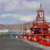 Un buque de Salvamento Marítimo anclado en el puerto de Arguineguín, en el sur de Gran Canaria. Alejado a la derecha, se encuentra una unidad móvil de la Cruz Roja Española. Al fondo, un complejo de edificios turísticos con un malecón y detrás, unas colinas