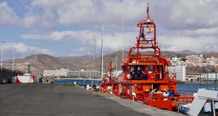 Un buque de Salvamento Marítimo anclado en el puerto de Arguineguín, en el sur de Gran Canaria. Alejado a la derecha, se encuentra una unidad móvil de la Cruz Roja Española. Al fondo, un complejo de edificios turísticos con un malecón y detrás, unas colinas