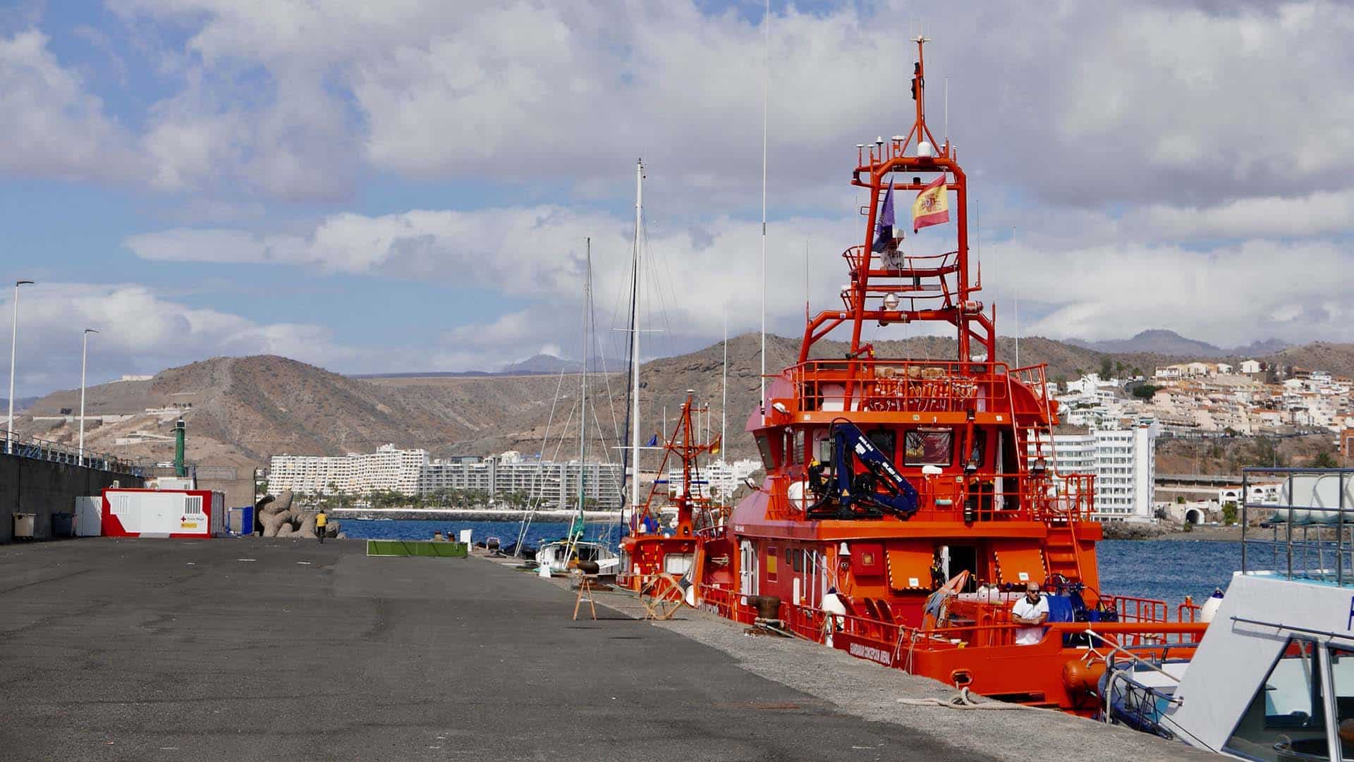 Un buque de Salvamento Marítimo anclado en el puerto de Arguineguín, en el sur de Gran Canaria. Alejado a la derecha, se encuentra una unidad móvil de la Cruz Roja Española. Al fondo, un complejo de edificios turísticos con un malecón y detrás, unas colinas