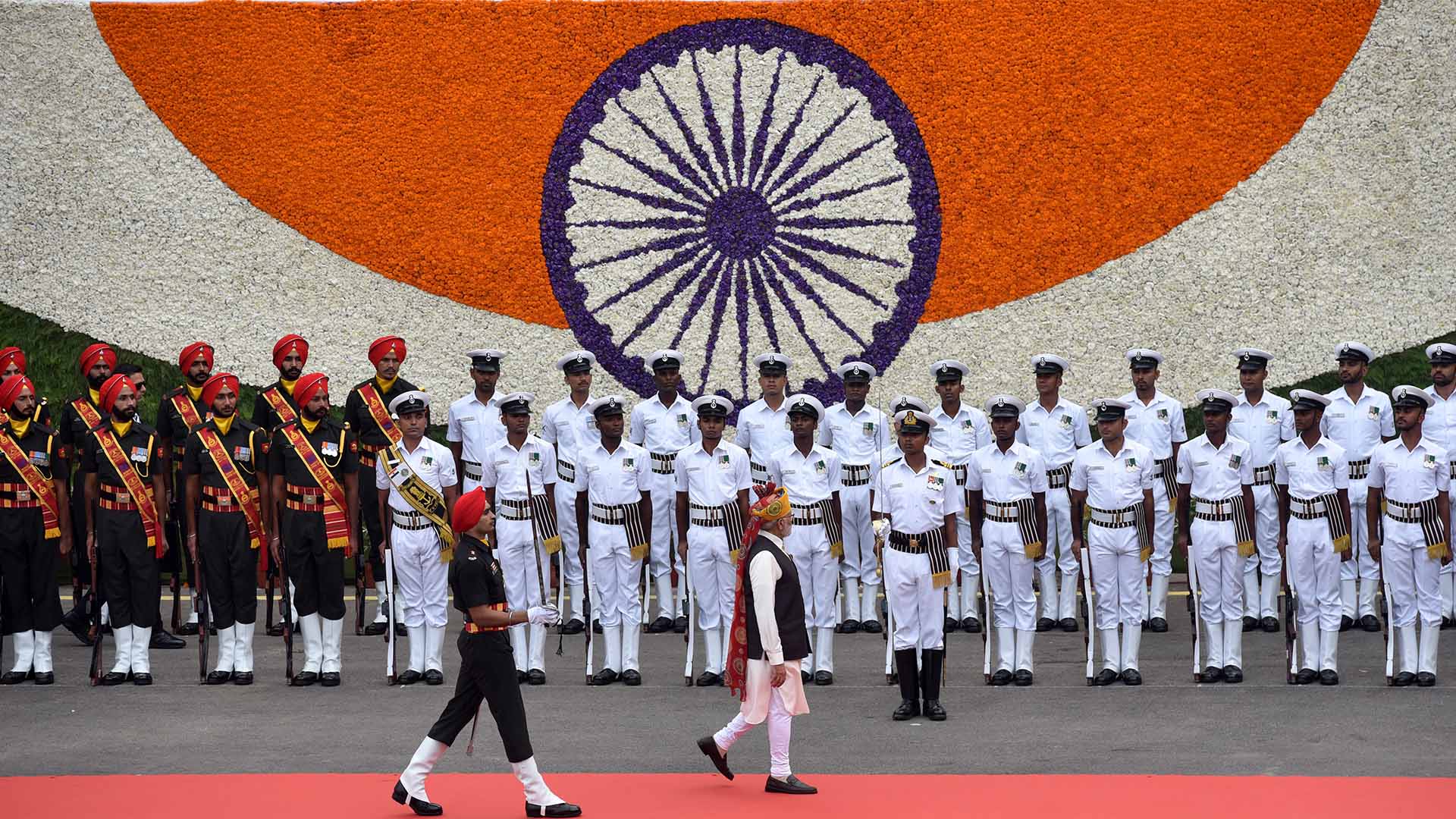 El primer ministro de la India, Narendra Modi, pasa revista a la Guardia de Honor durante el 77º acto del Día de la Independencia en el histórico Fuerte Rojo, el 15 de agosto de 2023 en Nueva Delhi. Modi es seguido por una guarda y al fondo un detalle de la bandera de India hecha con flores