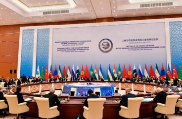 Plenario de la reunión de la Organización de Cooperación de Shanghái (OCS) en Samarcanda (Uzbekistán), en septiembre de 2022, con la participación de líderes de los Estados miembros sentados en una mesa redonda. Al fondo un panel de la reunión en los idiomas oficiales y las banderas con sus mástiles de los distintos países pertenencientes a la OCS