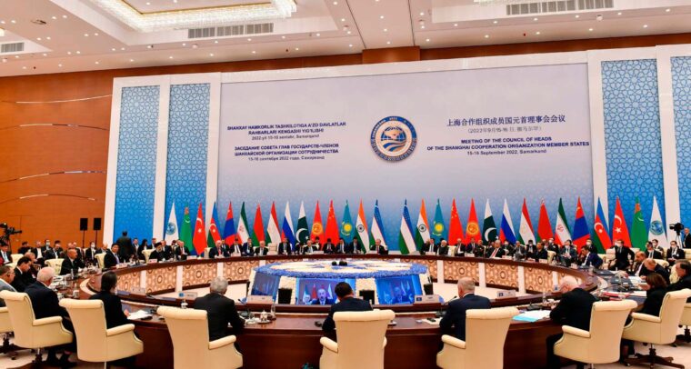 Plenario de la reunión de la Organización de Cooperación de Shanghái (OCS) en Samarcanda (Uzbekistán), en septiembre de 2022, con la participación de líderes de los Estados miembros sentados en una mesa redonda. Al fondo un panel de la reunión en los idiomas oficiales y las banderas con sus mástiles de los distintos países pertenencientes a la OCS