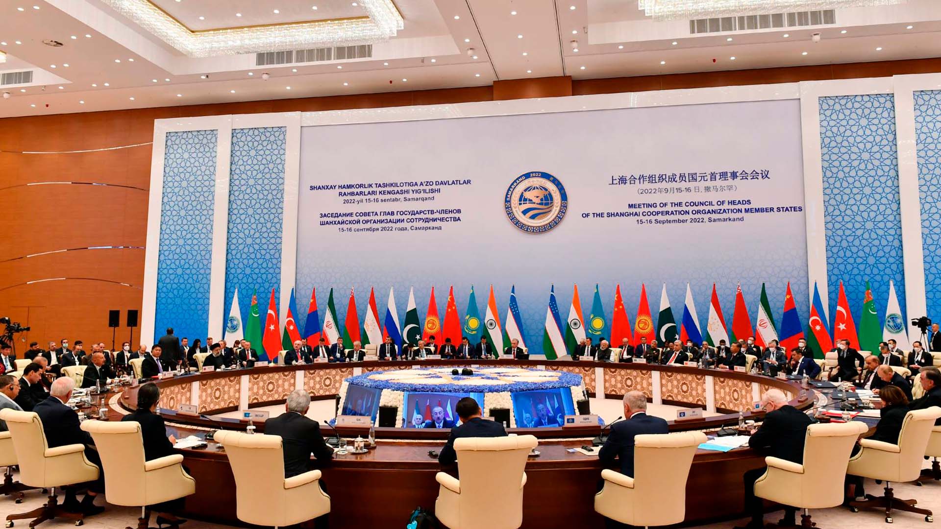 Plenario de la reunión de la Organización de Cooperación de Shanghái (OCS) en Samarcanda (Uzbekistán), en septiembre de 2022, con la participación de líderes de los Estados miembros sentados en una mesa redonda. Al fondo un panel de la reunión en los idiomas oficiales y las banderas con sus mástiles de los distintos países pertenencientes a la OCS