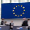 Imagen desenfocada de entre bastidores durante la sesión plenaria del Parlamento Europeo de octubre de 2024 en Estrasburgo (Francia). Al fondo, en una pantalla, la bandera de la UE.
