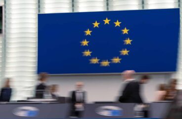 Imagen desenfocada de entre bastidores durante la sesión plenaria del Parlamento Europeo de octubre de 2024 en Estrasburgo (Francia). Al fondo, en una pantalla, la bandera de la UE.