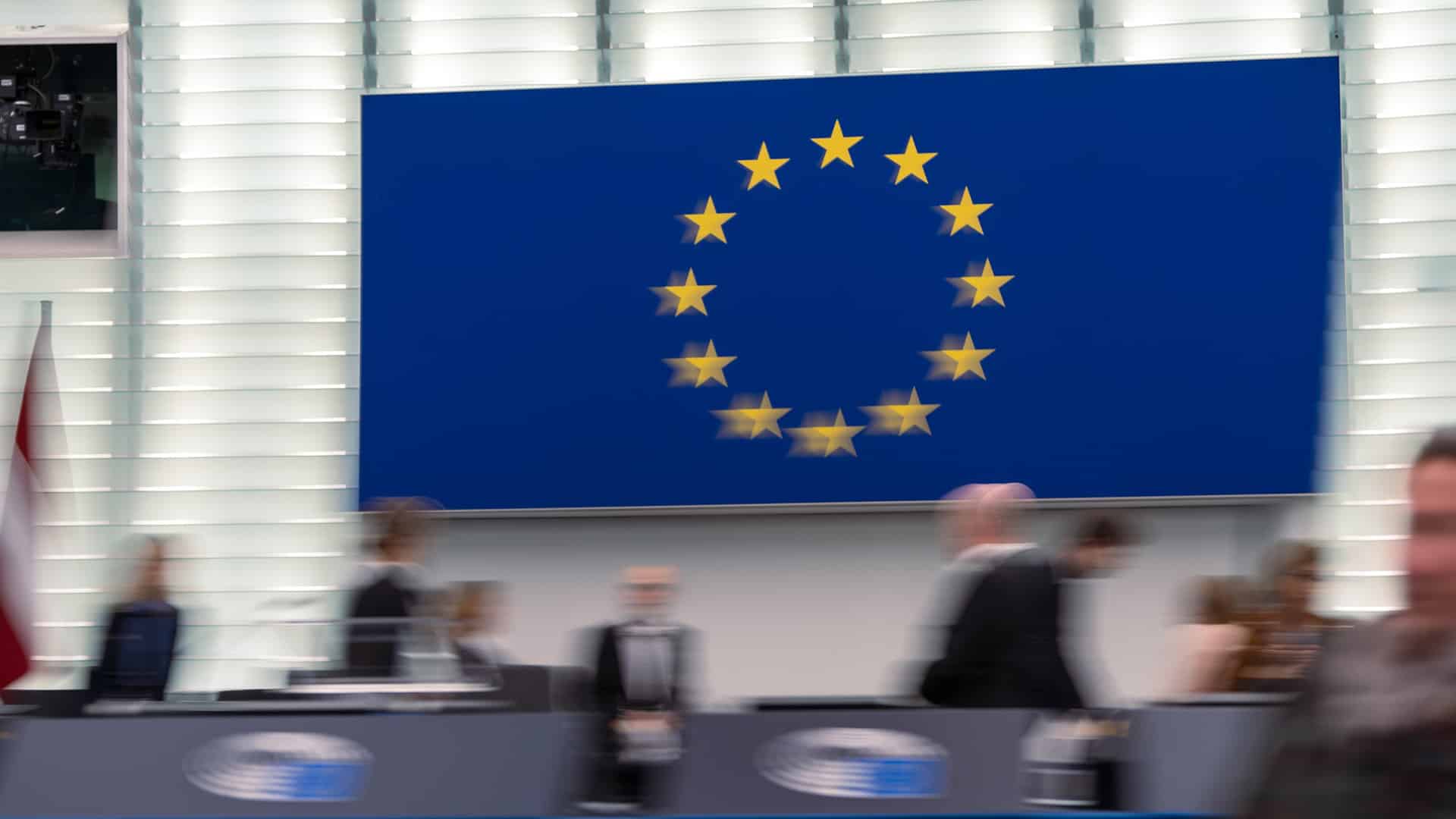 Imagen desenfocada de entre bastidores durante la sesión plenaria del Parlamento Europeo de octubre de 2024 en Estrasburgo (Francia). Al fondo, en una pantalla, la bandera de la UE.
