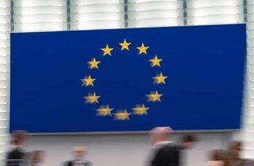 Session at the European Parliament, with the European Union logo in the background and blurred human figures in motion. Deepening for widening