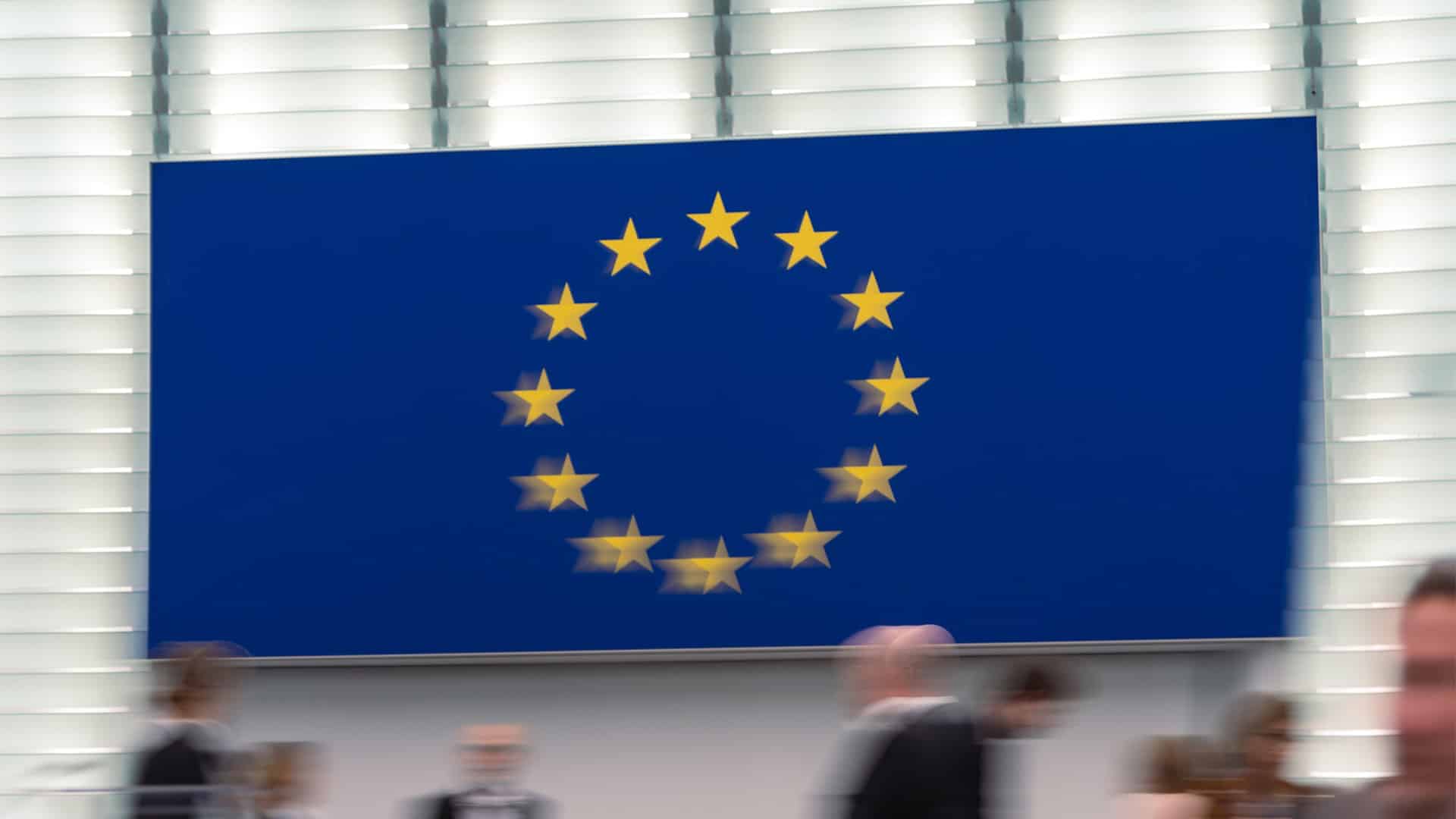 Sesión en el Parlamento Europeo, con el logotipo de la Unión Europea en el fondo y figuras humanas en movimiento desenfocado. Profundizar para ampliar