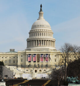 Frontal del edificio del Capitolio de los Estados Unidos, con estatuas y un estanque en primer plano, y banderas estadounidenses colgadas en la fachada principal, con la tribuna de juramento para la nueva Presidencia presente.