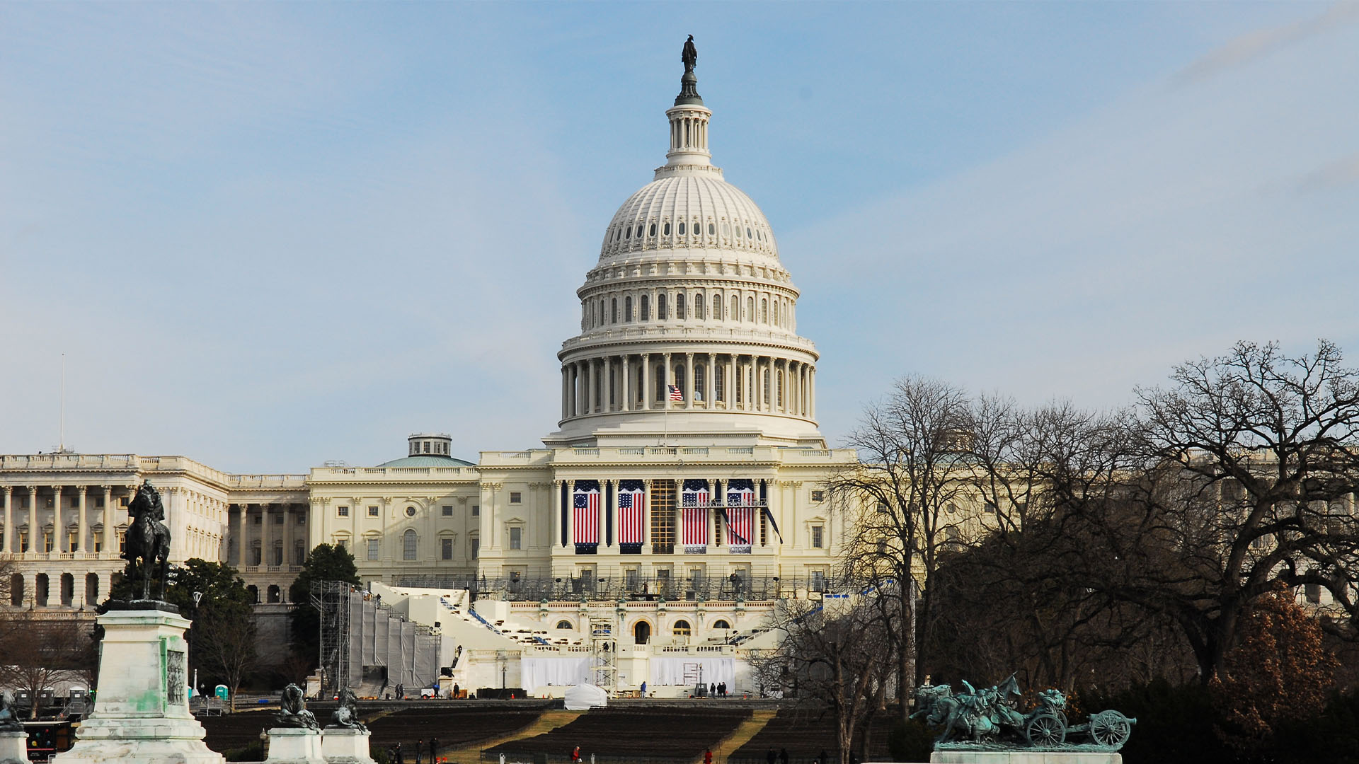 Frontal del edificio del Capitolio de los Estados Unidos, con estatuas y un estanque en primer plano, y banderas estadounidenses colgadas en la fachada principal, con la tribuna de juramento para la nueva Presidencia presente.