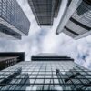 Vista hacia el cielo desde la base de varios rascacielos de vidrio y acero, reflejando las nubes en un día parcialmente nublado. La imagen representa la solidez y magnitud del sector financiero en un entorno urbano. Política fiscal