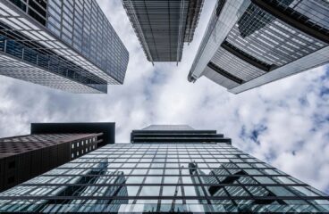 Vista hacia el cielo desde la base de varios rascacielos de vidrio y acero, reflejando las nubes en un día parcialmente nublado. La imagen representa la solidez y magnitud del sector financiero en un entorno urbano. Política fiscal