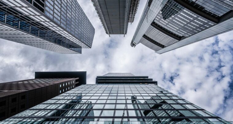 Vista hacia el cielo desde la base de varios rascacielos de vidrio y acero, reflejando las nubes en un día parcialmente nublado. La imagen representa la solidez y magnitud del sector financiero en un entorno urbano. Política fiscal