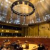 Audiencia en la Gran Sala del Tribunal de Justicia de la Unión Europea, con jueces sentados en un estrado curvo y abogados en el área central, bajo una gran estructura circular de iluminación. Inteligencia artificial