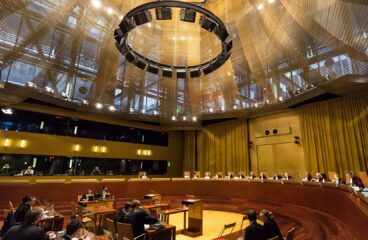 Audiencia en la Gran Sala del Tribunal de Justicia de la Unión Europea, con jueces sentados en un estrado curvo y abogados en el área central, bajo una gran estructura circular de iluminación. Inteligencia artificial